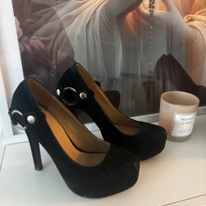 Svarta pumps med hög klack - Snygga svarta pumps med hög klack och rund tå. Skorna har en dekorativ rem med silverfärgad ring och knapp på sidan. Perfekta för att lyfta vilken outfit som helst med sin klassiska och stilrena look.