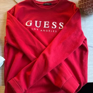Röd sweatshirt från Guess - Röd sweatshirt från Guess med vit logga framtill. Klassisk rund halsringning och långa ärmar. Mjuk bomullskvalitet och ribbade muddar vid ärmslut och nederkant. Perfekt för en chill och snygg look.