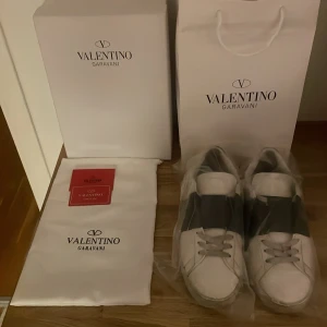 Valentino Garavani Open sneakers vit/svart - Säljer ett par Valentino Garavani Open sneakers i vitt skinn med bred svart rem över mitten. Klassisk rund tå, platt sula och snörning. Kommer med originalkartong, dustbag och äkthetskort. Riktigt snygga och stilrena sneakers för dig som gillar lyxiga detaljer.