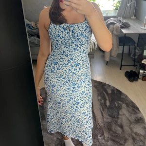 Blommig blåvit långklänning med knyt - Supersöt långklänning i vitt med blått blommönster och smala axelband. Klänningen har en feminin passform, knytning framtill vid bysten och är figurnära upptill. Perfekt för varma dagar och somriga vibes!