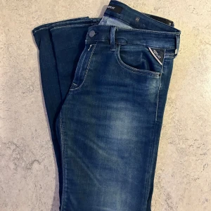 Blåa Replay Jeans Anbass - Snygga blå jeans från Replay i storlek W31 L30, med slim passform och klassisk femficksdesign. Jeansen är helt nya och har en mörk tvätt med lätt slitning och tydliga Replay-detaljer vid fickan. Perfekt för dig som gillar en stilren look.