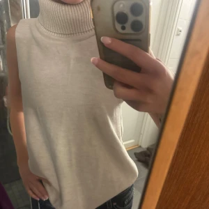 Beige ärmlös polotopp - Säljer en oandvänd stilren beige polotopp utan ärmar. Toppen har en hög, ribbad polokrage och är i ett mjukt, stickat material. Perfekt för lager-på-lager eller som den är till jeans. Enkel och clean look som passar till mycket.