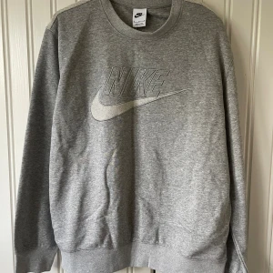 Grå sweatshirt från Nike  - Grå sweatshirt från Nike i mjuk bomullsmix med broderad Nike-logo framtill. Rund halsringning och långa ärmar med ribbade muddar. 