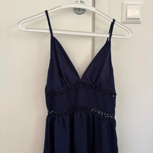 Marinblå sommarklänning med spetsdetaljer - Snygg marinblå klänning med smala axelband och v-ringning. Klänningen har dekorativa spetsdetaljer under bysten och i midjan, vilket ger en extra touch. Perfekt för varma dagar och har en luftig känsla.