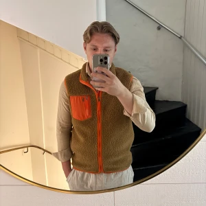 Brun fleeceväst från Polo Ralph Lauren - Säljer en brun fleeceväst från Polo Ralph Lauren med orange detaljer och dragkedja. Västen har en orange bröstficka med dragkedja och kontrasterande orange kantband. Perfekt för lager-på-lager och riktigt snygg till streetwear.