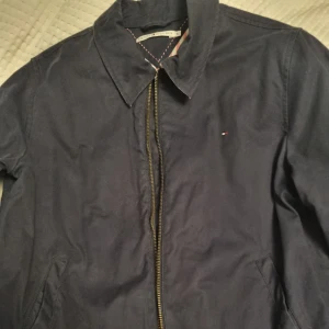 Mörkblå jacka Tommy Hilfiger  - Snygg mörkblå vindjacka från Tommy Hilfiger i klassisk modell med dragkedja framtill och diskret logga på bröstet. Jackan har krage, sidfickor och randigt innerfoder vid halsen. Står XL men är mer som L
