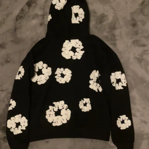 Svart hoodie med vita blommor Denim Tears - Svart hoodie från Denim Tears i storlek M med stora vita blommönster över hela tröjan, inklusive huvan. Tröjan har en klassisk passform, ribbade muddar och huva. Perfekt för dig som gillar streetwear och vill sticka ut med ett unikt mönster.
