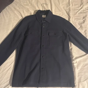 Mörkblå overshirt från Selected Homme - Snygg mörkblå overshirt från Selected Homme med klassisk krage, två bröstfickor med lock och knappar framtill. Perfekt lager-på-lager-plagg med stilren look och lång ärm. Materialet är mjukt och känns riktigt skönt mot huden. Den är även helt oanvänd då den inte passar mig!