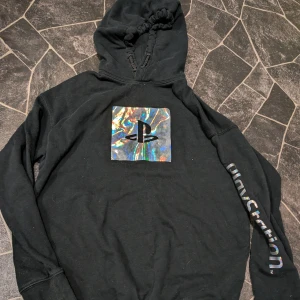 Svart PlayStation hoodie med holografiskt tryck stl 146-152 - Svart hoodie från PlayStation med ett holografiskt tryck på bröstet och PlayStation-logga på ena ärmen. Tröjan har huva med snörning och är tillverkad i mjuk bomull och polyesterblandning. Perfekt för dig som gillar gaming och vill ha en chill stil.