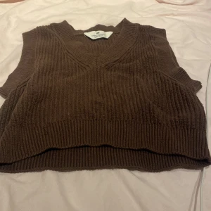 Brun stickad väst från H&M - Mysig brun stickad väst från H&M med v-ringning och ribbad struktur. Perfekt att slänga över en skjorta eller t-shirt för en trendig lager-på-lager-look. Västen har en något croppad passform och breda muddar vid hals och nederkant.