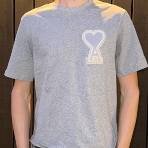Grå t-shirt från Ami Paris med hjärtdetalj - Snygg grå t-shirt från Ami Paris med ett stort broderat hjärta och A-logga på bröstet. Klassisk rund halsringning och korta ärmar. Tillverkad i mjuk bomull som känns skön mot huden. Perfekt för en clean och stilren look.