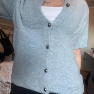 Ljusblå kofta från Gina Tricot - Supermjuk ljusblå kofta från Gina Tricot med korta ärmar och snygga silvriga knappar framtill. Koftan är stickad och har en klassisk ribbad kant vid halsen och ärmsluten. Perfekt att slänga över en topp för en chill look. Storlek M men passar S också!💞