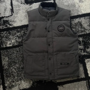 Väst - Snygg mörkgrå dunväst från Cahobc Goose med hög krage och raka axlar. Västen har två stora fickor framtill, knäppning med svarta knappar och ett broderat märke med texten 'Arctic Parker' på bröstet. Perfekt för kyliga dagar och enkel att styla över en hoodie. Passar för folk mellan 170-180,  den är stor i storlek 