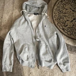 Grå hoodie från Polo Ralph Lauren - Säljer en grå hoodie från Polo Ralph Lauren med dragkedja och huva med vita snören. Klassisk logga broderad på bröstet och två fickor framtill. Perfekt för chill dagar eller när du vill ha en enkel och stilren look. DET STÅR XXL MEN KÄNNS SOM M (Har storlek M) skriv vid frågor! 🤔🧑🏽‍🤝‍🧑🏽🤝🏽 Den är i gott skick förutom att tjedjan gått (ur led) men det kan man fixa inom 10 min hemma👌🏽