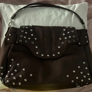 Brun axelväska med nitar - Cool brun axelväska från Carin Wester i syntetmaterial med silverfärgade nitar på locket och hörnen. Väskan har ett brett lock med tryckknapp och ett rymligt huvudfack. Perfekt för dig som gillar detaljer och vill sticka ut lite extra.