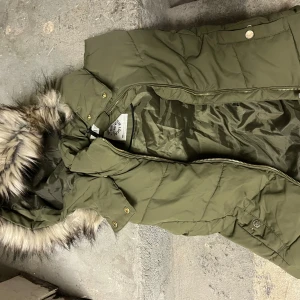 Olivgrön pufferväst från H&M - Säljer en olivgrön pufferväst från H&M med fuskpäls på huvan och guldfärgade detaljer. Västen har dragkedja och tryckknappar framtill, quiltad design och är perfekt för kalla dagar. Snygg och praktisk med cool look.