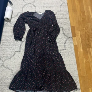 Svart långklänning med småblommigt mönster - Supersöt svart långklänning från Gina Tricot med små röda blommor, v-ringning och långa ärmar med volangdetalj vid ärmslut. Klänningen har omlottkänsla och markerad midja, perfekt för dig som gillar romantisk stil.