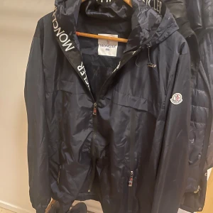 Svart vindjacka från Moncler  - Säljer en vindjacka från Moncler i storlek XXL dock känns den mer som en XL. Jackan har huva med Moncler-logga, dragkedja framtill och flera fickor med dragkedja. Materialet är glansig polyester och jackan har en cool serietidningsdetalj på insidan.