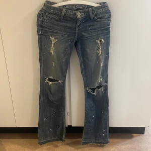 Blå slitna bootcut jeans från Guess  - Säljer ett par blå jeans från Guess med bootcut passform. Jeansen har slitna detaljer, hål vid knäna och råa benslut. Färgskiftningar och stänk ger en cool, vintage vibe. Klassisk femficksmodell med Guess-logga på bakfickan och insidan av linningen.