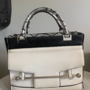 Vit och svart handväska med ormmönster - Snygg handväska i vit och svart med ormmönstrade detaljer på handtag och kanter. Väskan har en strukturerad yta som liknar reptilskinn, silverfärgade metalldetaljer och en avtagbar axelrem. Perfekt för dig som gillar statement-accessoarer och coola detaljer.