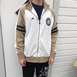 Nike track jacket beige och vit - Nike track jacket i beige och vitt med svarta ränder på ärmarna och broderad logga på bröstet. Jackan har hög krage, hel dragkedja och muddar vid ärmslut och nederkant. Perfekt för en sportig och avslappnad stil.