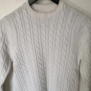 Vit kabelstickad tröja från 157 - Snygg vit kabelstickad tröja från 157 med rund hals och ribbade muddar. Tröjan har ett klassiskt flätmönster och är gjord i mjuk bomull, perfekt för att hålla dig varm under kyliga dagar. Passar dig som gillar stilrena och tidlösa plagg.