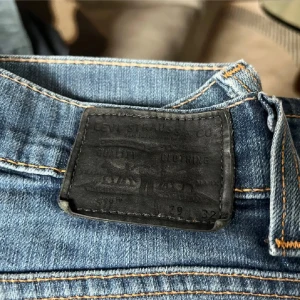 Levi's 511 blå jeans - Säljer ett par klassiska Levi's 511 jeans i blå denim med snygg slitning och orange sömmar. Modellen har normal passform och raka ben, med fem fickor och svart läderpatch bak i midjan. Perfekta för dig som gillar en tidlös jeanslook.