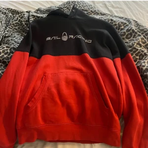Röd och svart hoodie från Sail Racing - Säljer en snygg hoodie från Sail Racing i rött och svart med logga framtill. Tröjan har huva, magficka och är tillverkad i mjuk bomull. Perfekt för dig som gillar streetwear och vill ha något bekvämt och stilrent.