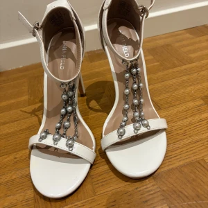 Vita klackade sandaler med pärldetaljer - Snygga vita sandaler med hög klack från Anna Field. Skorna har öppen tå, smal rem över tårna och dekorativa kedjor med pärlor framtill. Stängs med spänne runt ankeln. Perfekta för dig som vill sticka ut med lite extra bling. Använt ett par gånger