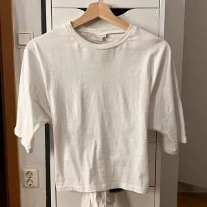 Vit kortärmad topp med knyt från Nelly - Vit kortärmad topp från Nelly i mjuk bomull. Toppen har rund halsringning och en snygg knyt-detalj i ryggen som ger en trendig look. Perfekt till jeans eller kjol för en clean och fräsch stil. Inga defekter!