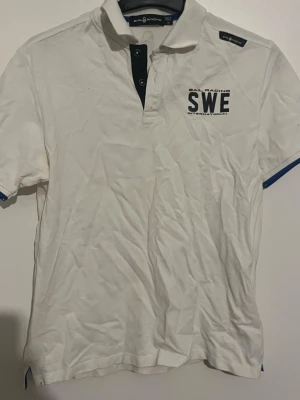 Vit piké från Sail Racing - Vit pikétröja från Sail Racing med trycket 'SWE' på bröstet. Tröjan har korta ärmar, knappslå med mörkblå detaljer och blå kantband vid ärmsluten. Perfekt för en sportig och avslappnad stil. Storlek xs (170) barnstorlek