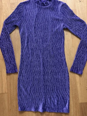 Lila långärmad kortklänning med öppen rygg - Säljer en lila kortklänning med lång ärm och coolt mönster. Klänningen har en tight passform och en stor öppning i ryggen som ger en snygg detalj. Perfekt för dig som vill sticka ut med en unik look.