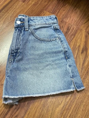 Blå denimskort med rå kant - Trendigt blått denimskort från AMY med fransig, rå kant och klassiska fickor både fram och bak. Skorten har en asymmetrisk design framtill och är tillverkad i 100% bomull. Perfekt för en avslappnad och cool stil.
