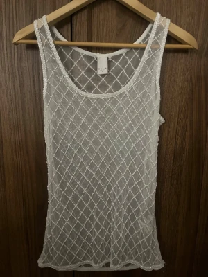 Vit meshlinne från Vila - Säljer ett vitt meshlinne från Vila med ett snyggt rutmönster av pärlor. Linnet är transparent och har breda axelband samt rund halsringning. Perfekt att styla över ett linne eller bikini för en trendig look.