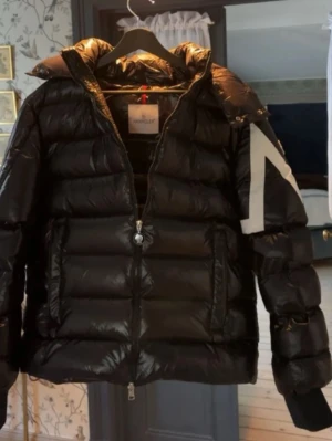 Svart dunjacka från Moncler - Snygg svart dunjacka från Moncler med glansig finish och vit detalj på ärmen samt Moncler-logga. Jackan har dragkedja framtill, hög krage och vadderad design som håller dig varm under kalla dagar.