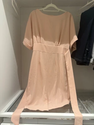 Ljusrosa klänning med knytskärp - Söt ljusrosa klänning med korta ärmar och rund halsringning. Klänningen har ett brett knytskärp i midjan som ger en fin siluett. Modellen är rak och enkel, perfekt för en stilren look. Materialet känns mjukt och lätt.