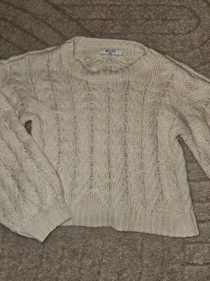 Beige kabelstickad tröja från NA-KD - Snygg beige kabelstickad tröja från NA-KD med rund halsringning och långa ärmar. Tröjan har ett klassiskt flätmönster och ribbade muddar vid ärmslut och nederkant. Perfekt för lager-på-lager och en chill look.