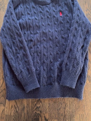 Mörkblå kabelstickad tröja Polo Ralph Lauren - Mörkblå kabelstickad tröja från Polo Ralph Lauren med röd broderad logga på bröstet. Tröjan har rund halsringning, långa ärmar och ribbstickade muddar. Perfekt för lager-på-lager och klassisk stil.