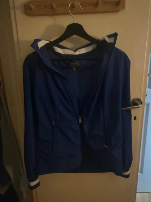 Raffe windbreaker storlek 14-16 - Snygg blå vindjacka från Polo Ralph Lauren med vit kant på huvan och ärmsluten. Jackan har dragkedja framtill, två fickor med dragkedja och en luva. Perfekt för blåsiga dagar och enkel att matcha med det mesta.