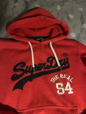 Röd vintage Superdry hoodie - Röd hoodie från Superdry med stor logga och texten 'The Real 54' broderat på bröstet. Klassisk känguruficka framtill, vita dragsnören och mjukt material. Perfekt för dig som gillar sportig och avslappnad stil.