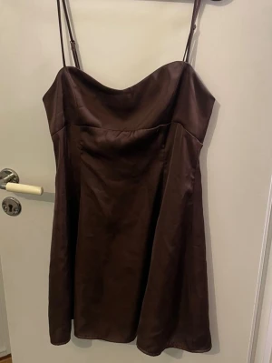Brun kort klänning med tunna axelband - Snygg brun kort klänning med tunna justerbara axelband och lätt utsvängd passform. Klänningen har en slät framsida och smockad rygg för extra komfort. Perfekt för en trendig och stilren look.