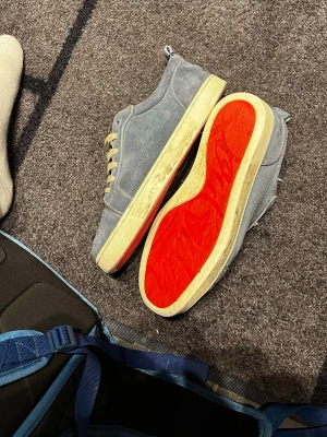 Cristian Louboutin  - Snygga gråa sneakers i mocka med beige snören och innersula. Skorna har en ikonisk röd yttersula och beige mellansula. Perfekta för dig som vill sticka ut med en unik detalj på skorna. Klassisk låg modell med rund tå och diskret design.