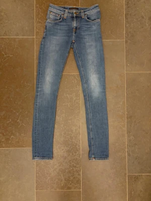 Blå slim fit jeans från Nudie Jeans - Snygga blå slim fit jeans från Nudie Jeans med klassisk femficksdesign och subtila slitningar framtill. Jeansen har normal midja och smala ben, perfekta för en avslappnad men trendig look. Materialet är mjukt och stretchigt för extra komfort.