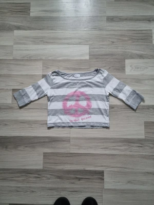 Randig croppad topp med peace-tryck - Croppad topp med breda grå och vita ränder, trekvartsärm och stort rosa peace-märke framtill. Texten 'Do what you love!' under trycket. Perfekt för en avslappnad och cool stil.