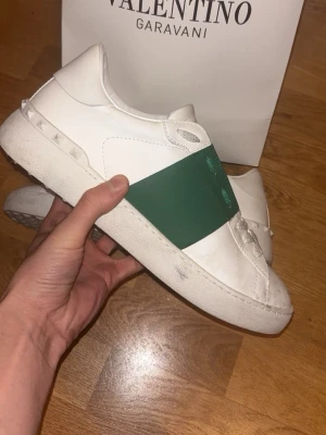 Valentino open sneakers gröna - Snygga vita sneakers från Valentino med en bred grön rem över mitten. Skorna har klassisk låg profil, vita nitar på hälen och är tillverkade i skinn. Perfekta för dig som vill ha en stilren men ändå unik look. Påse ingår och lite andra små saker men box ingår inte. 