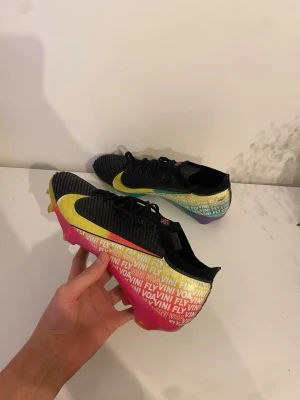 Nike Mercurial fotbollsskor svart/grön/röd - Säljer ett par Nike Mercurial fotbollsskor med svart ovandel, stor gul swoosh och färgglad sula i gult, rosa och grönt. Texten 'FLY VINI' syns på hälen. Skorna har snörning och är tillverkade i syntetmaterial för bästa grepp och känsla på planen.