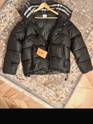 Burberry jacka - Snygg svart dunjacka från Burberry med stor vadderad huva och klassiskt rutigt foder. Jackan har dragkedja och tryckknappar framtill, två stora fickor och justerbar nederkant. Perfekt för kalla vinterdagar och riktigt trendig look. Priset kan diskuteras!!