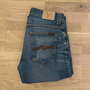 Blå jeans från Nudie Jeans - Säljer ett par klassiska blå jeans från Nudie Jeans med raka ben och snygga slitningar. Jeansen har kontrastsömmar och den ikoniska Nudie-bågen på bakfickorna. Perfekta för en avslappnad och trendig look.