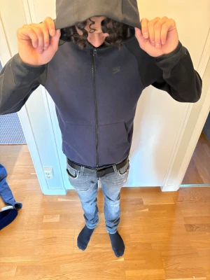 Mörkblå Nike hoodie med dragkedja - Snygg mörkblå hoodie från Nike med svarta ärmar och diskret Nike-logga på bröstet. Hoodien har dragkedja hela vägen, två fickor framtill och en stor huva. Perfekt för en chill och sportig look. Som kommer med byxorna 