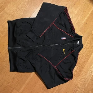 Svart Miami Heat vindjacka Nike NBA - Svart vindjacka från Nike med Miami Heat-tryck på ryggen och NBA-logga framtill. Jackan har röda detaljer längs ärmarna och gul Nike Swoosh på bröstet. Lätt material, dragkedja och ribbade muddar. Perfekt för basketfans som vill sticka ut.